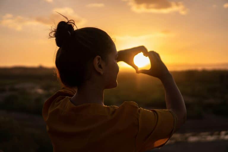 Une femme fait un coeur avec ses mains au soleil couchant